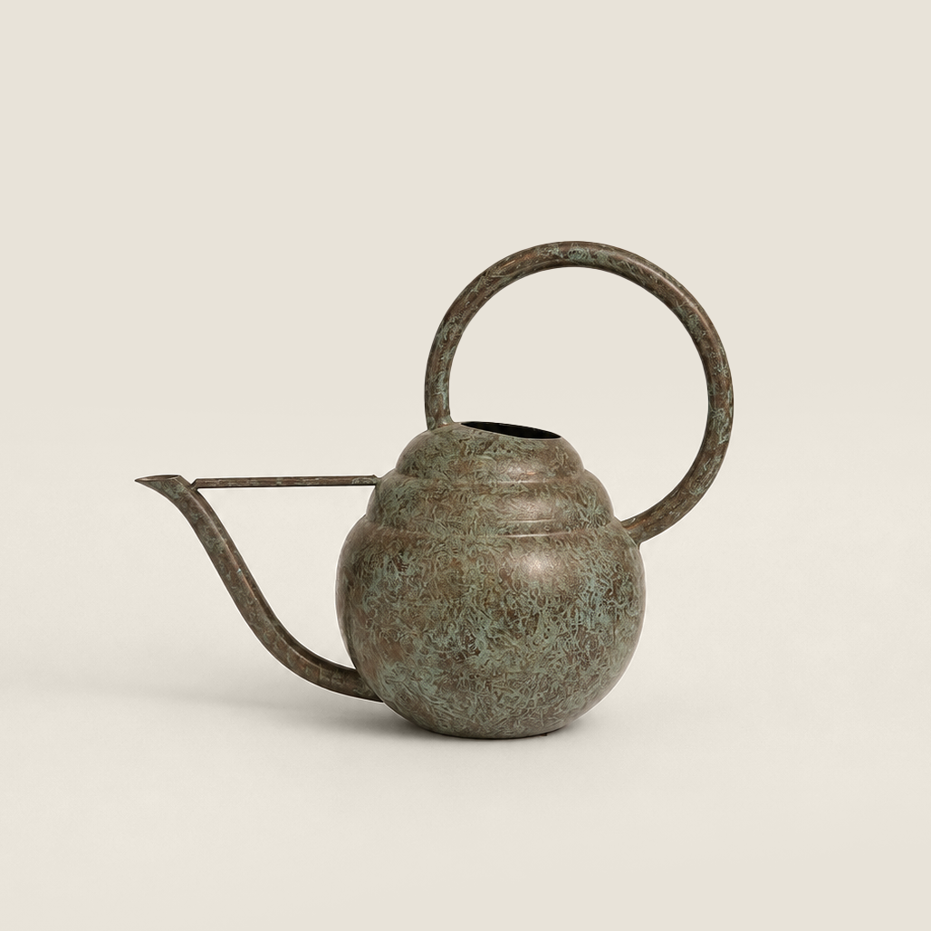 A small metal watering can with a rounded body, loop handle, and spout, finished in an aged bronze or verdigris-like metallic finish, photographed on a plain light warm-gray background.