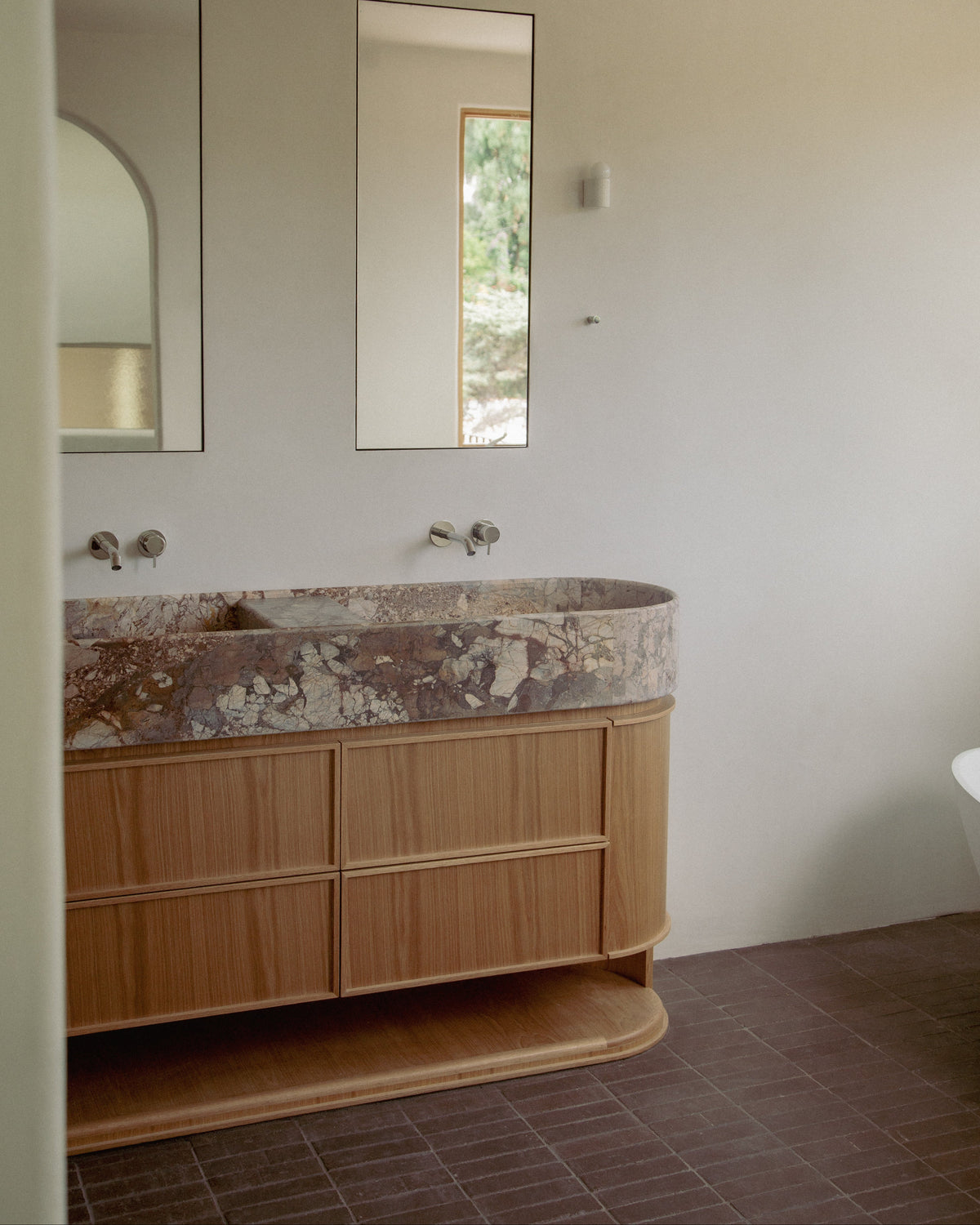 A modern bathroom vanity with a curved double-sink basin set into a light wood cabinet, featuring a textured stone countertop and minimalist wall mirrors above.
