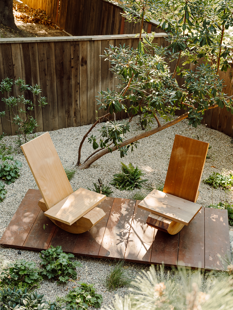 Wooden chairs on a wooden platform in a garden setting with plants and gravel.