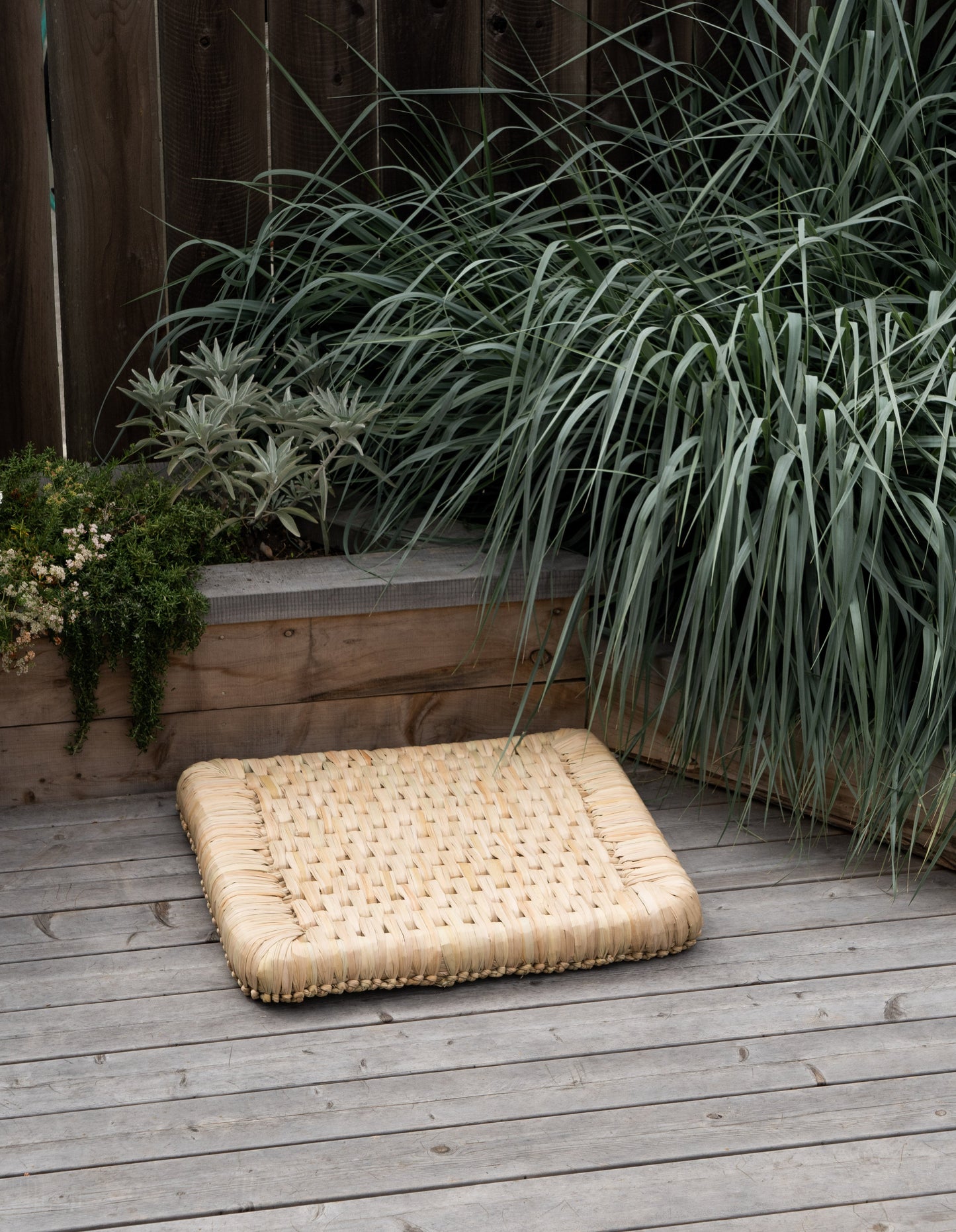 Woven mat on a wooden deck with plants and a wooden fence in the background