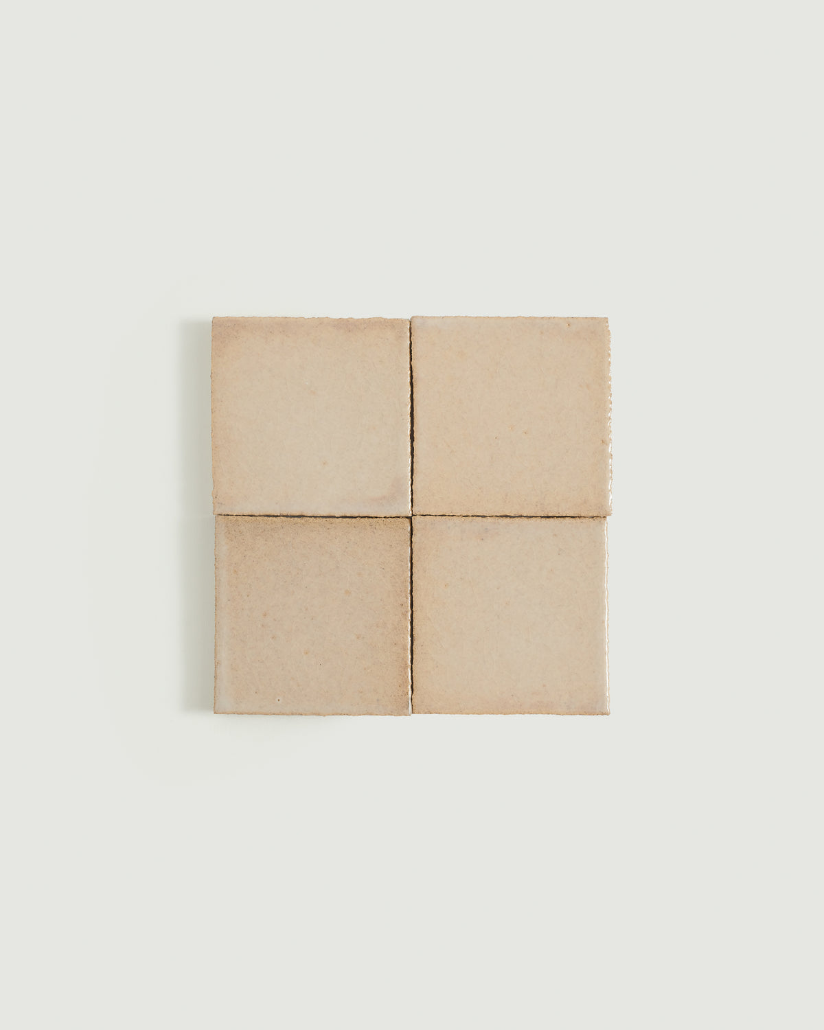 Four square ceramic tiles arranged in a 2x2 grid, featuring a matte, warm sandy beige finish with subtle texture and fine edge irregularities, set against a solid off-white background.