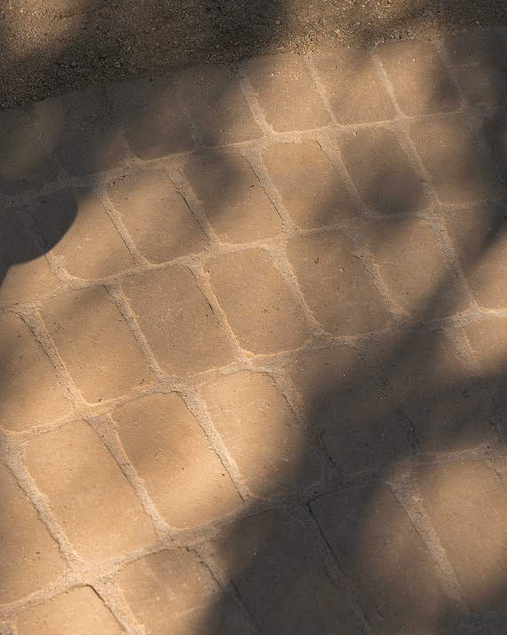 A close-up view of tan stone pavers with irregular rectangular shapes and light grout lines, cast in dappled sunlight and shadow.