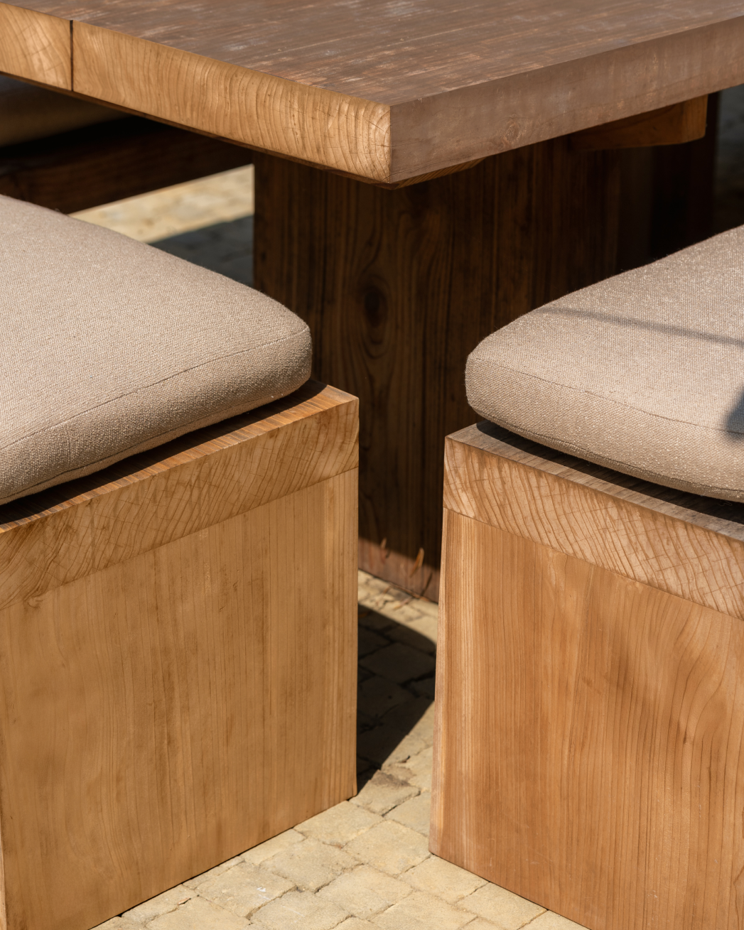 Outdoor wooden Montecito dining set with a thick-topped table and two matching bench seats with light beige cushions, shown on a paved surface in bright daylight.