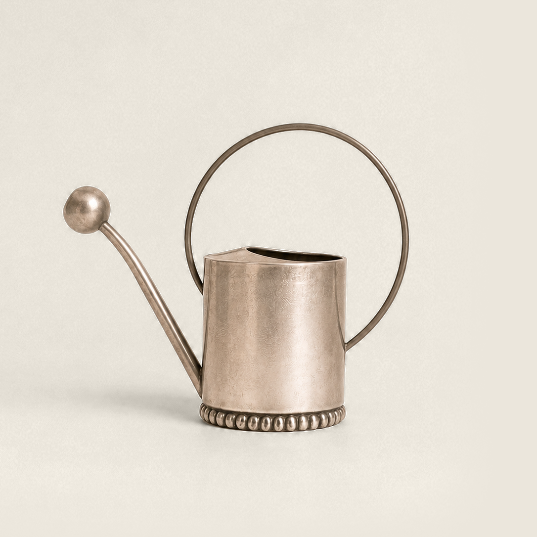 A small metallic watering can with a long spout, loop handle, and beaded base, photographed on a plain warm off-white background.