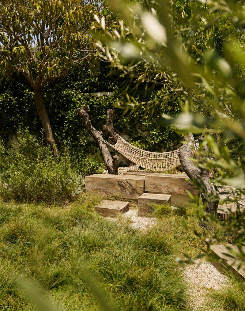 A rustic woven rope hammock suspended between two tree trunks in a lush green outdoor garden setting with stone steps and foliage.
