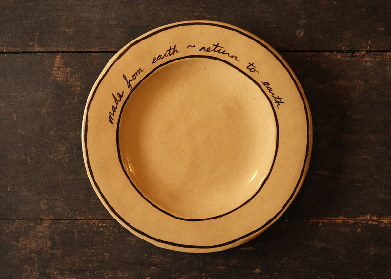 Beige ceramic plate with "made from earth, return to earth" inscribed text on a dark wooden surface