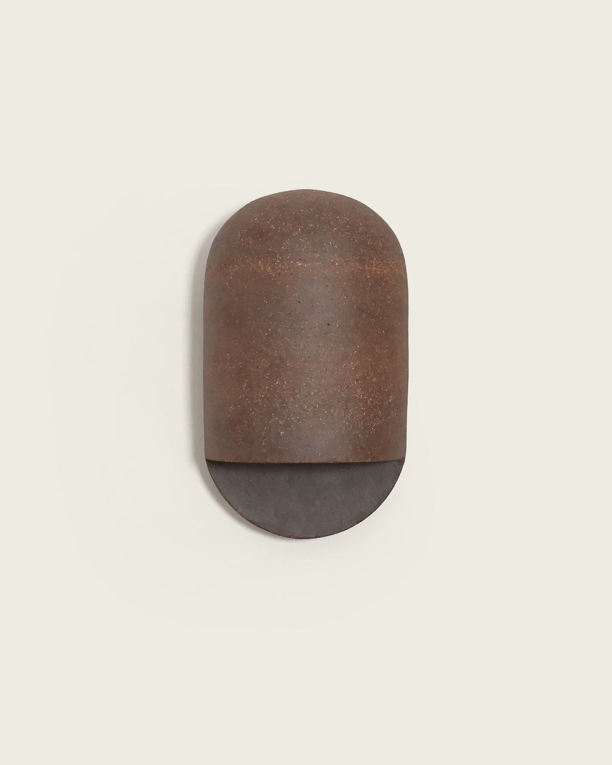 A minimalist wall-mounted light fixture with a rounded rectangular cap-like upper section in textured rust-brown metal and a semi-circular lower section in dark charcoal gray, shown against a solid light beige background.