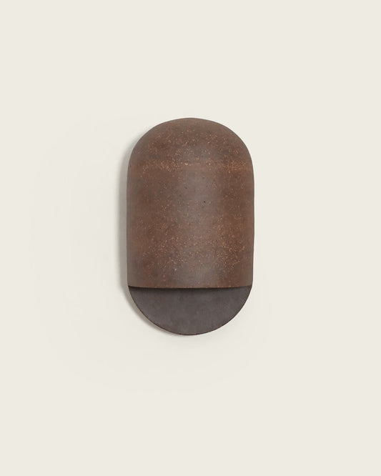 A minimalist wall-mounted light fixture with a rounded rectangular cap-like upper section in textured rust-brown metal and a semi-circular lower section in dark charcoal gray, shown against a solid light beige background.