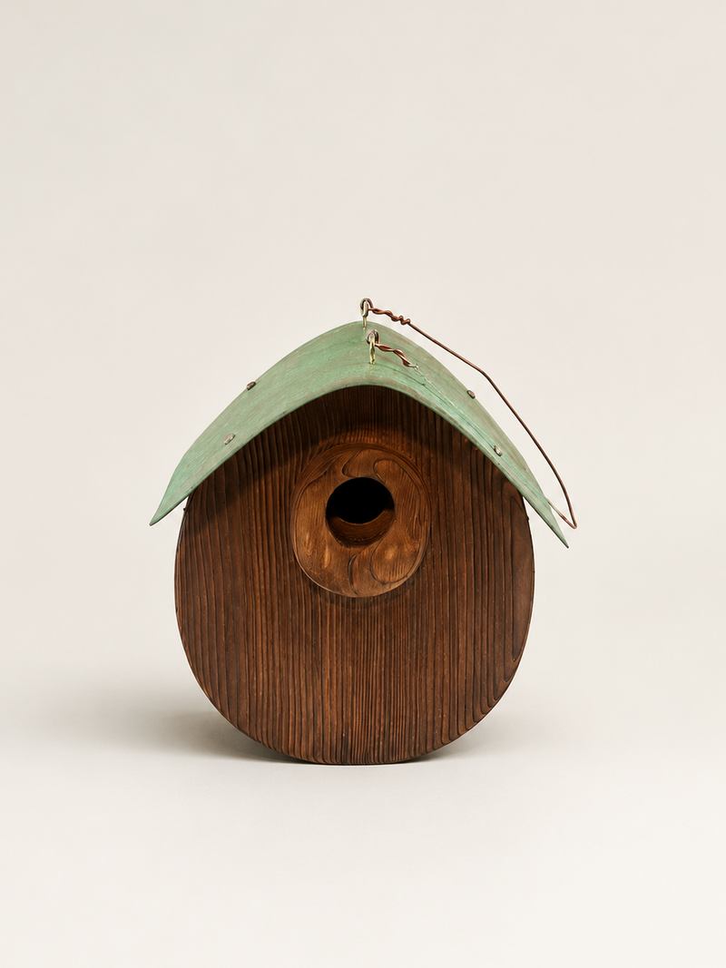 A rustic wooden birdhouse with a dark brown wood-grain body and a weathered light green metal roof, featuring a small hanging wire loop at the top.