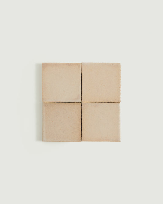 Four square ceramic tiles arranged in a 2x2 grid, featuring a matte, warm sandy beige finish with subtle texture and fine edge irregularities, set against a solid off-white background.