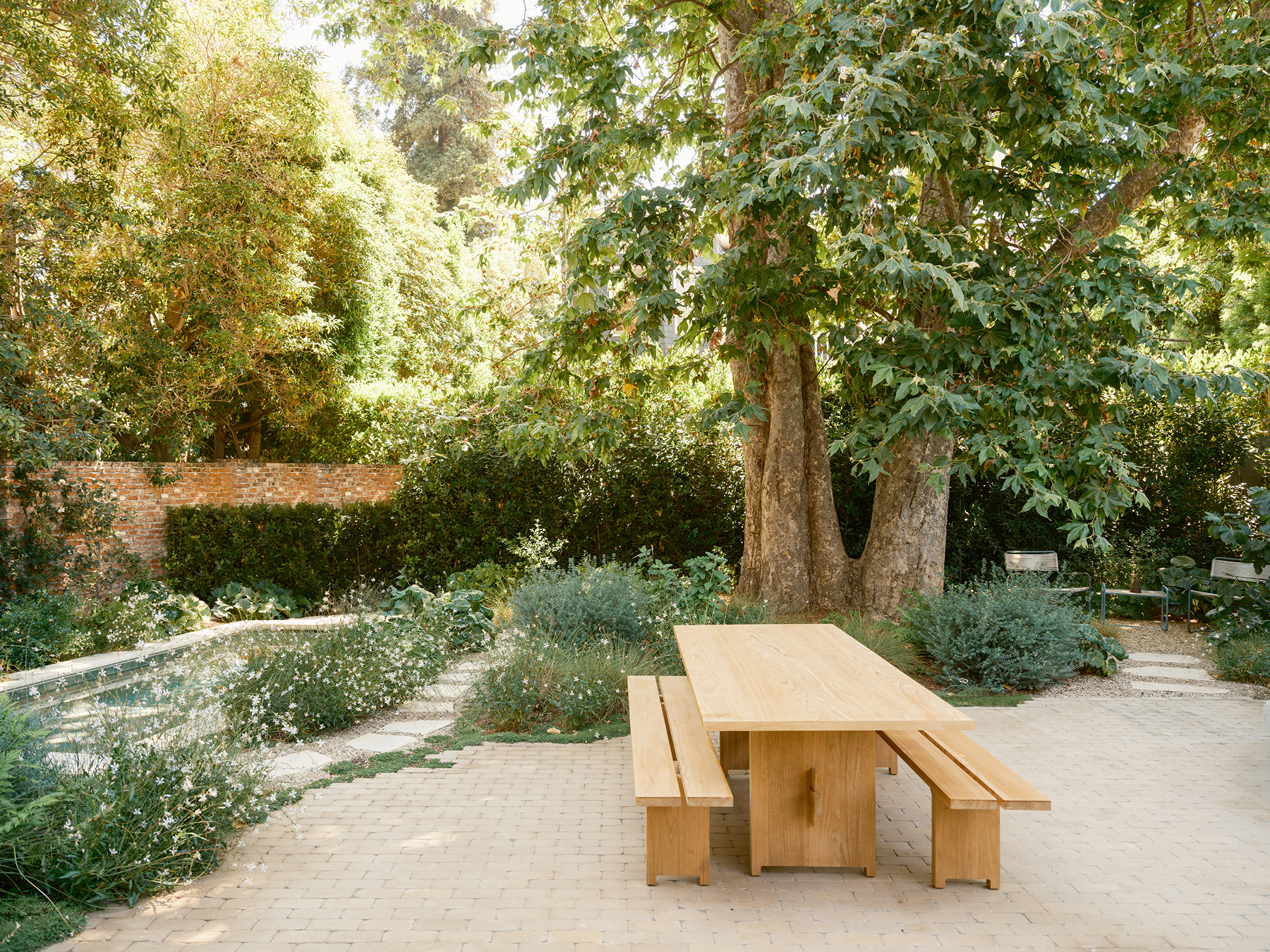 Outdoor wood dining table and benches in a garden setting with trees and greenery.