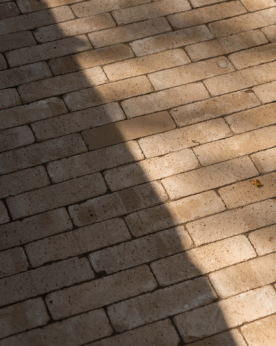 A close-up view of tan rectangular paving stones arranged in a brick pattern, with natural sunlight casting diagonal shadows across the surface.