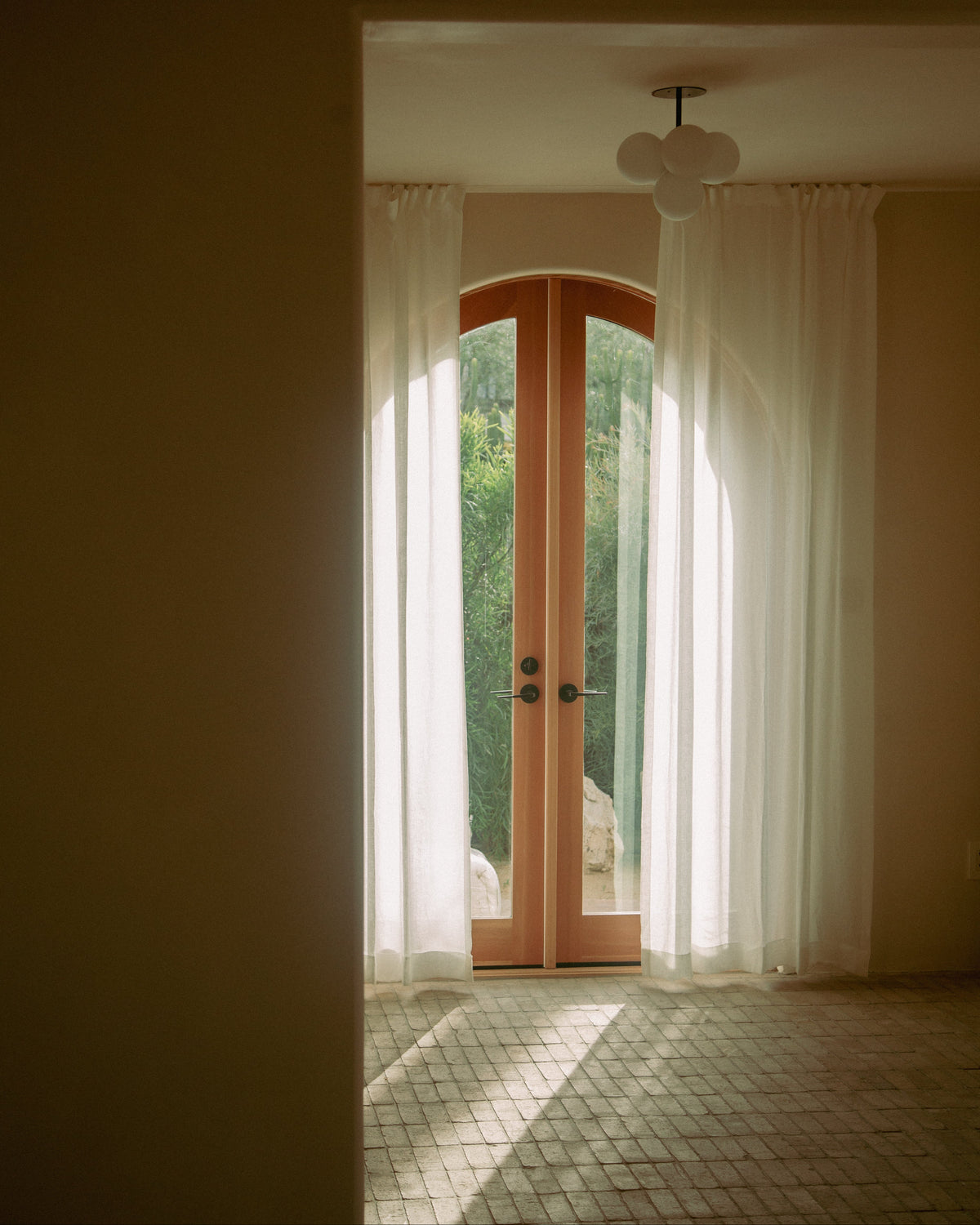 A view through a doorway into a sunlit room with sheer white curtains framing a pair of warm wood double doors, with natural light streaming in and a tiled floor below.