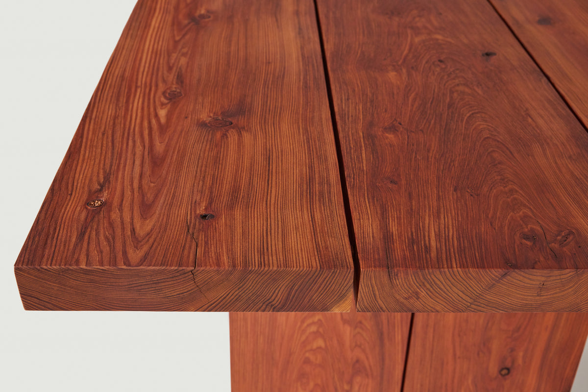 Close-up of a wooden table with thick slatted top and matching legs, showing rich reddish-brown wood grain and natural knots against a light neutral background.