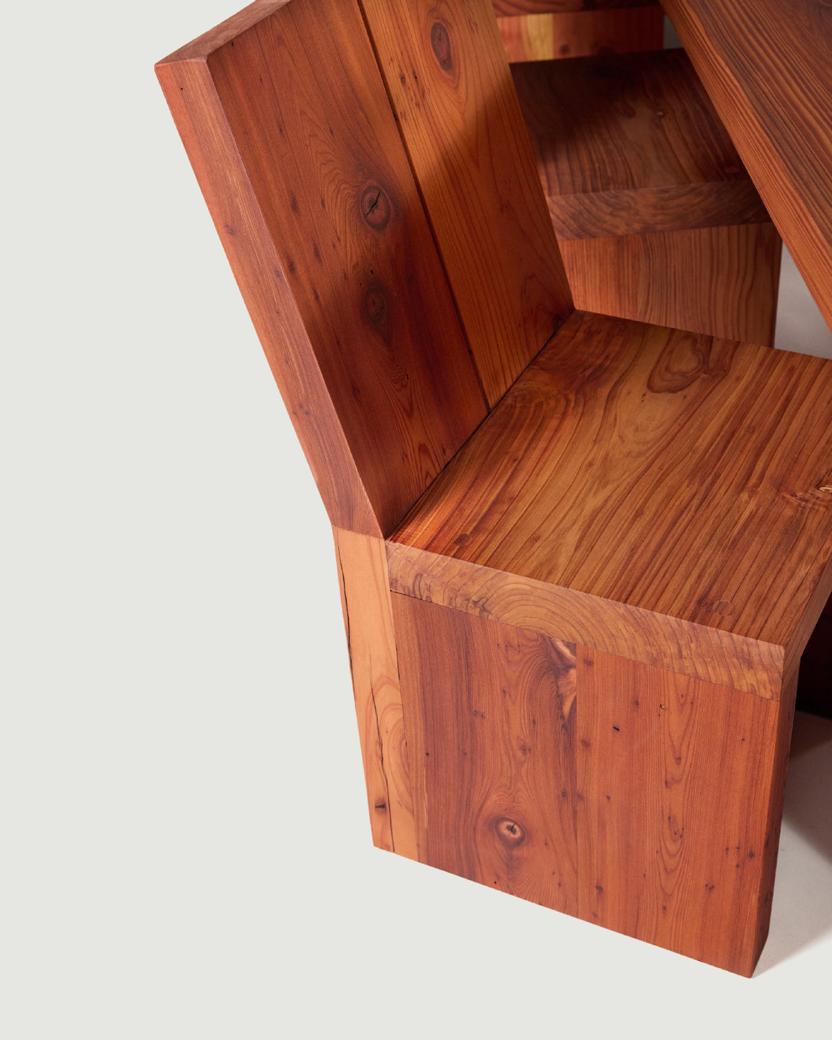 A close-up of a wooden chair with a thick blocky seat and back, showcasing warm reddish-brown wood grain and knots, set against a plain light gray background.