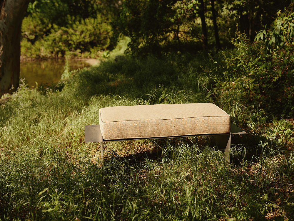 Metal bench with cream cushion in a grassy outdoor setting with trees and water in the background