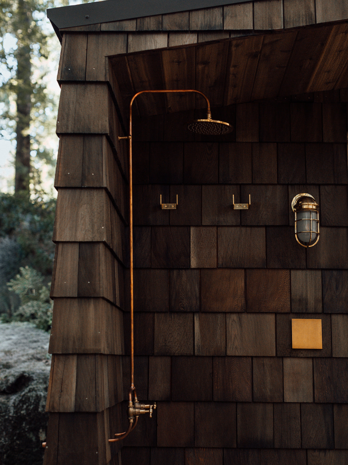 A outdoor shower setup with a brass shower and round shower head mounted on a dark wood shingled wall, with matching brass hooks and a light fixture nearby.
