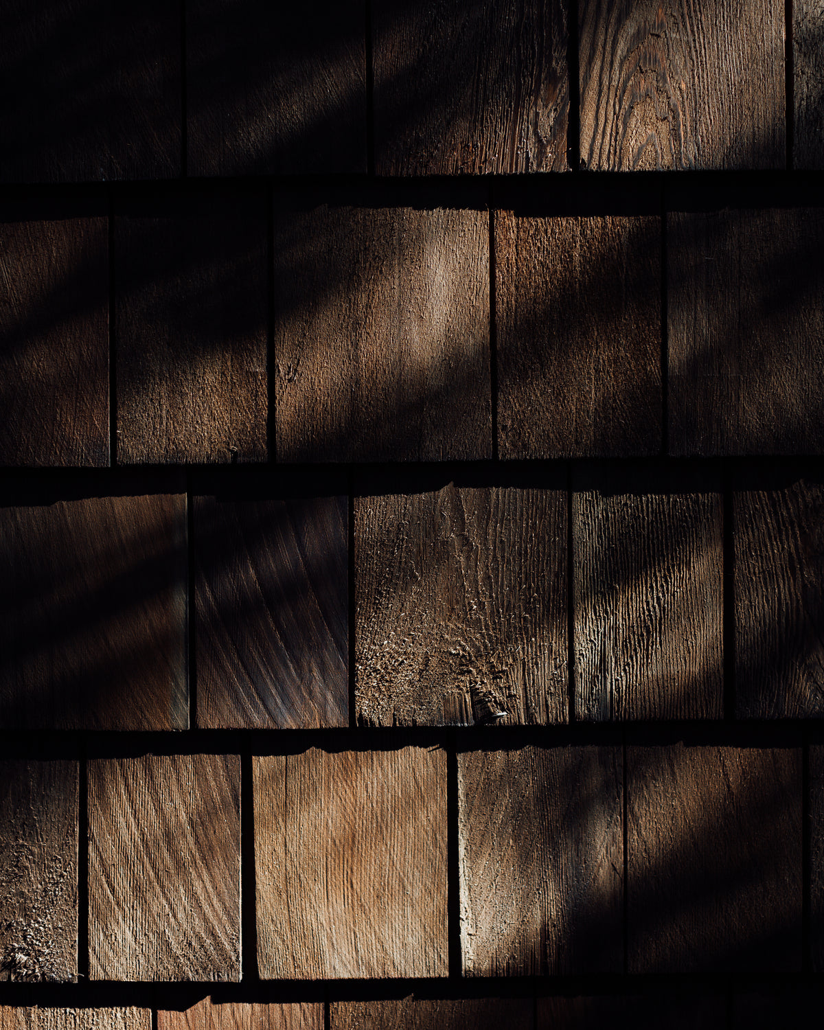 Dark brown wooden shingles with dramatic leaf shadow patterns cast across the textured surface.