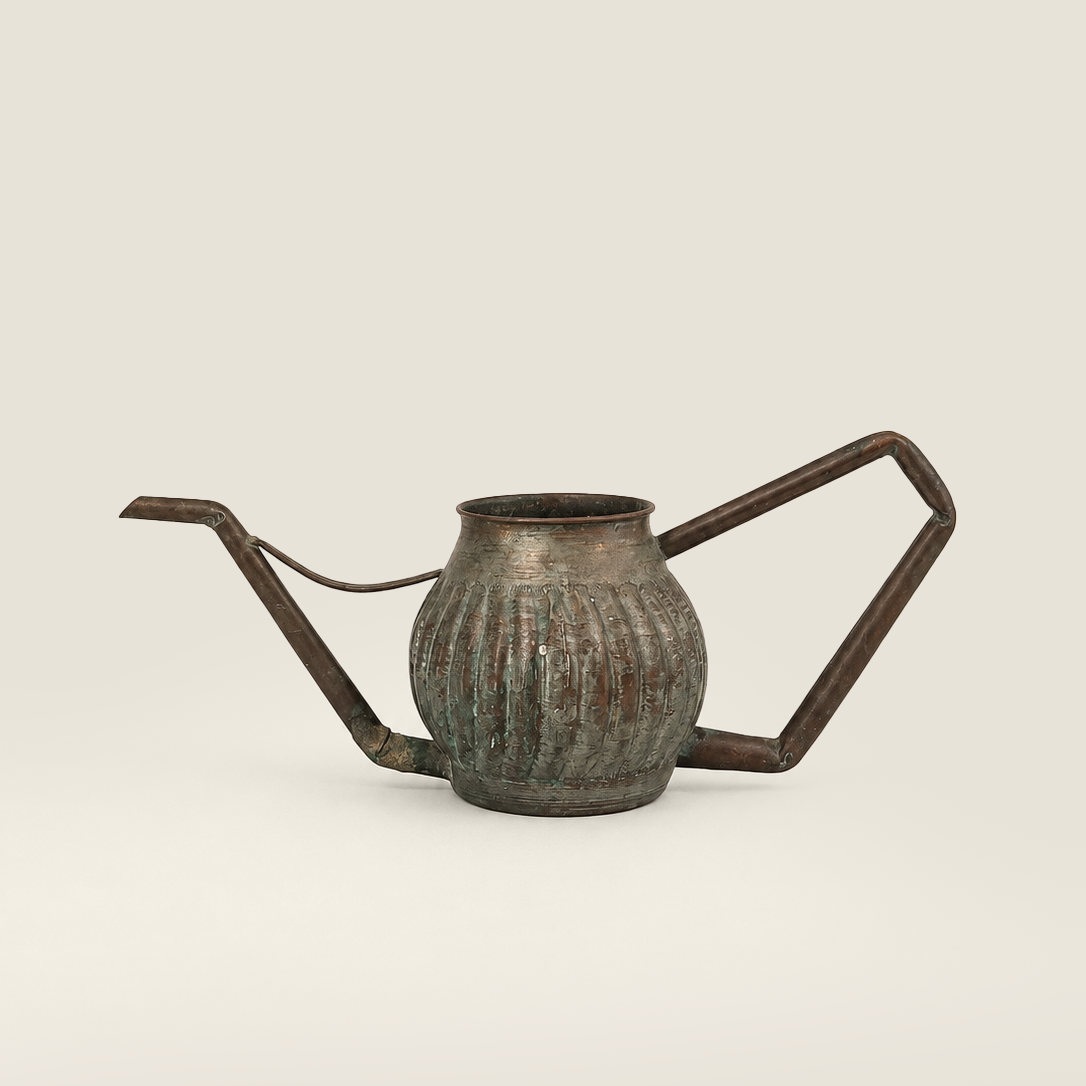 A vintage-style metal watering can a rounded ribbed body, a long angled spout, and a geometric side handle, shown on a plain light beige background.