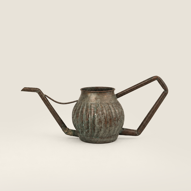 A vintage-style metal watering can a rounded ribbed body, a long angled spout, and a geometric side handle, shown on a plain light beige background.