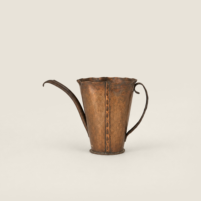 A vintage hammered metal pitcher with a long curved spout, a side loop handle, and a vertical seam with decorative stitching, shown on a solid light beige background.